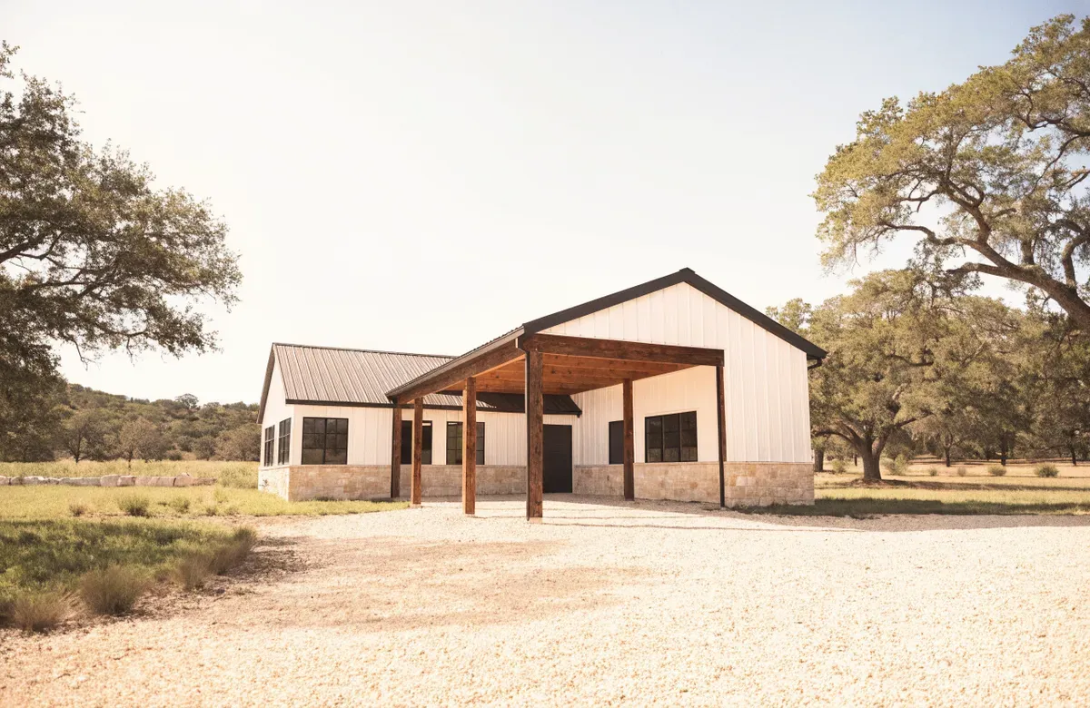 A white barndominium with limestone wainscot and a tall cedar-post covered entry under live oaks in the Texas Hill Country