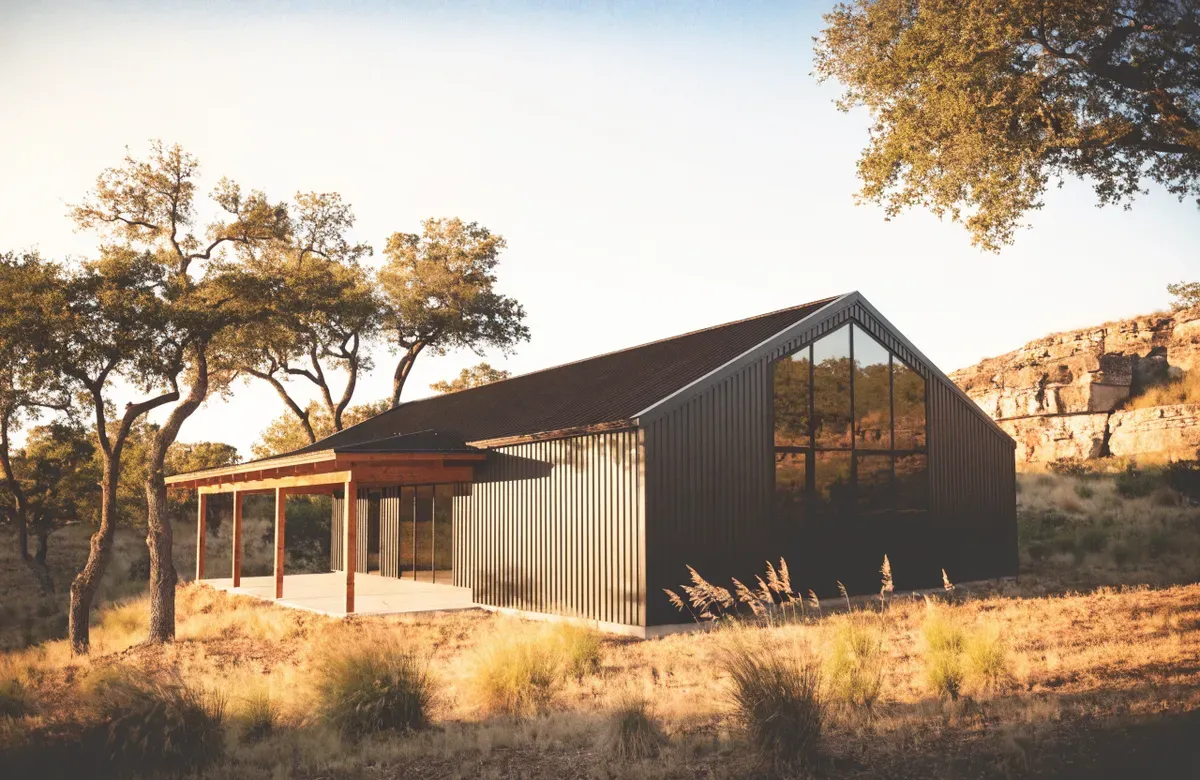 A charcoal metal barndominium with a large glass gable end and a covered patio beside a limestone bluff in the Texas Hill Country