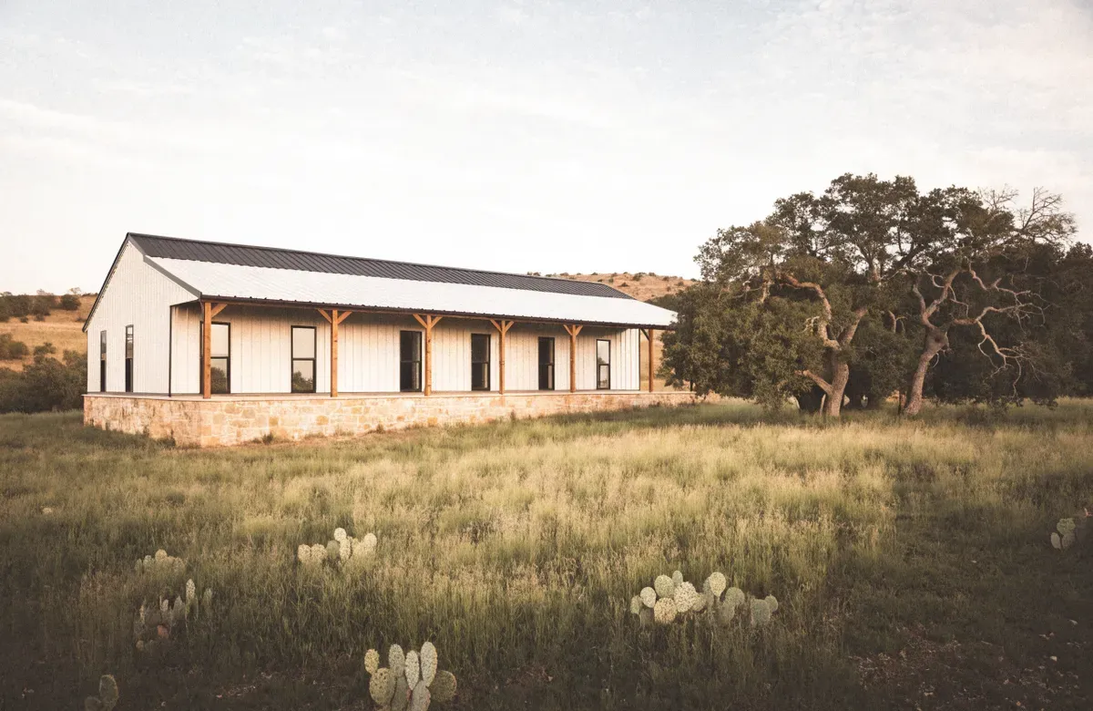 A long low barndominium with a limestone base, white metal siding, and a wide porch on Hill Country pasture with prickly pear