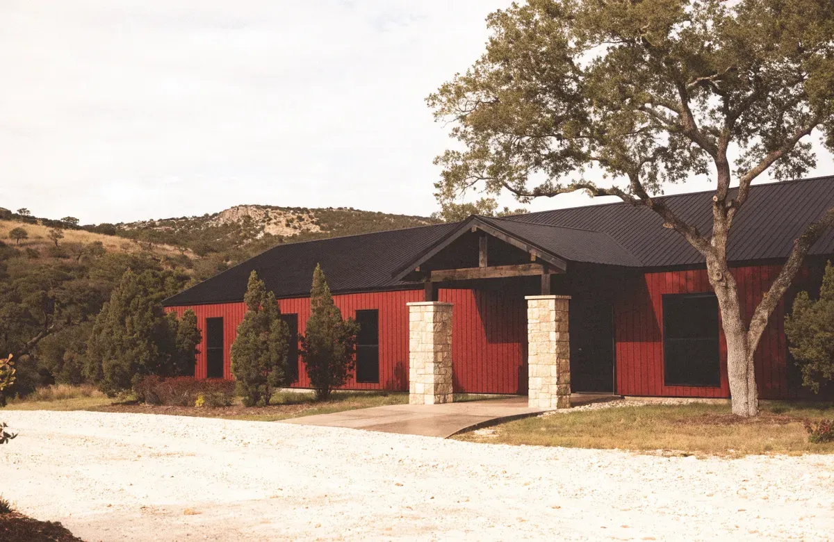 A red metal barndominium with limestone entry columns and a black standing-seam roof on wooded Hill Country acreage
