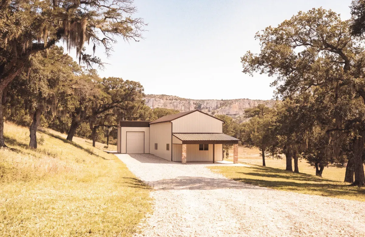 A cream barndominium with a tall attached RV and equipment bay and a covered porch below a Hill Country limestone bluff