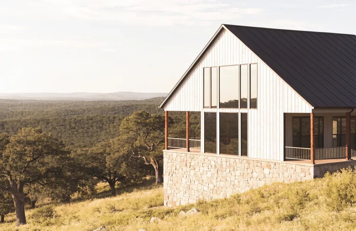 A two-story barndominium with a limestone lower level, white board-and-batten upper level, and a black metal roof above a Hill Country valley