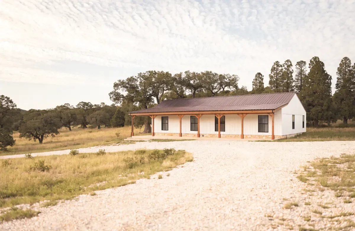 A single-story white barndominium with a bronze standing-seam roof and a full-length covered porch on Hill Country ranch land