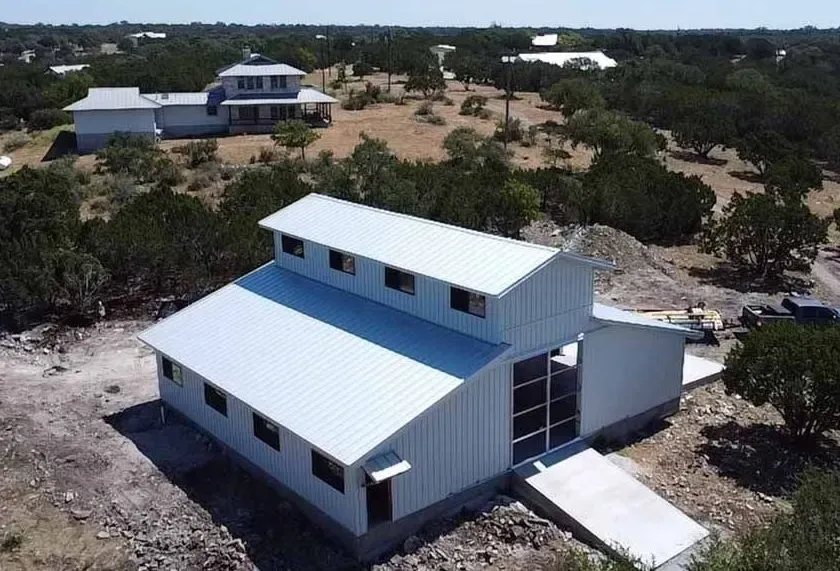 Aerial view of a light gray steel barndominium with a raised monitor roof and clerestory windows on a hillside lot in Texas