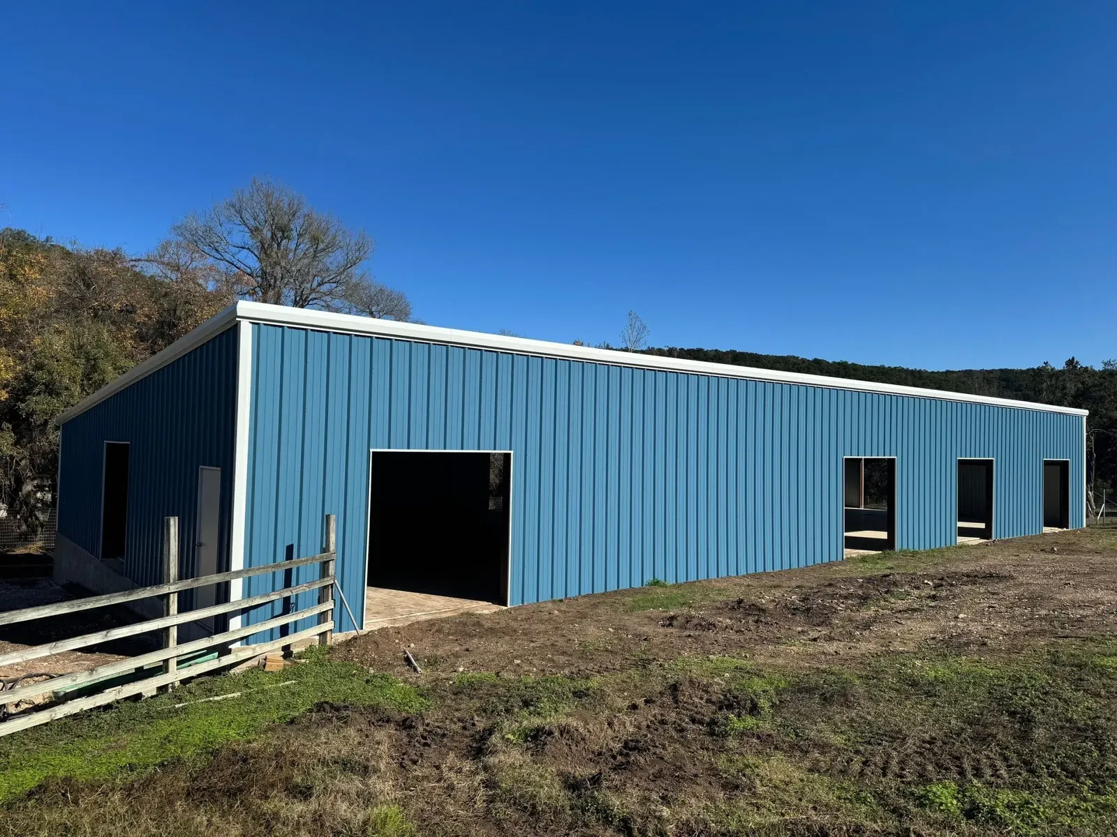A long blue steel agricultural building with several open bays behind a rail fence in Texas