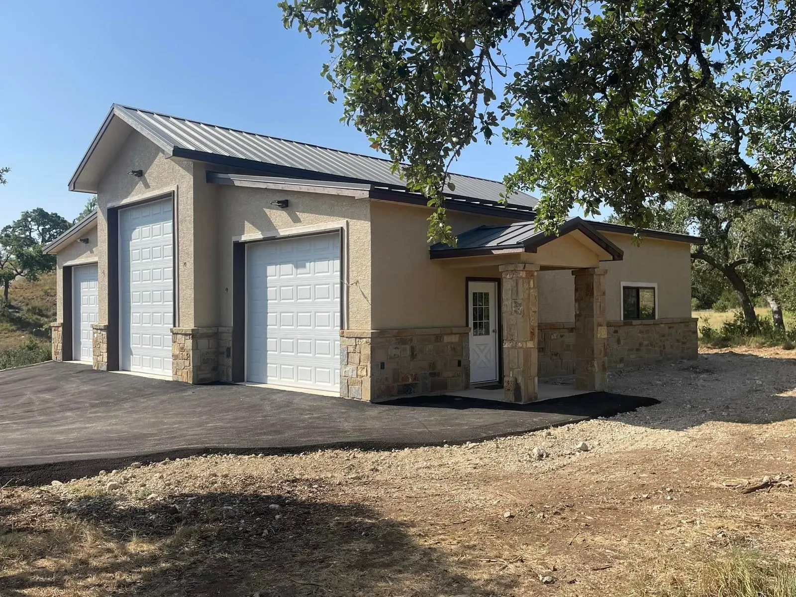 A stucco and stone three-bay steel garage with a covered stone entry under oak trees in Texas