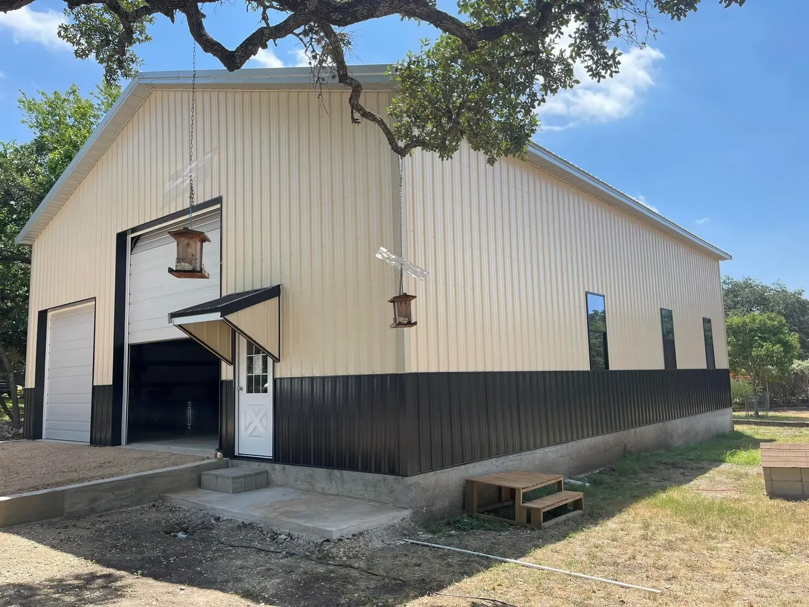A tan steel shop with black wainscot and a tall roll-up door under a live oak in Texas