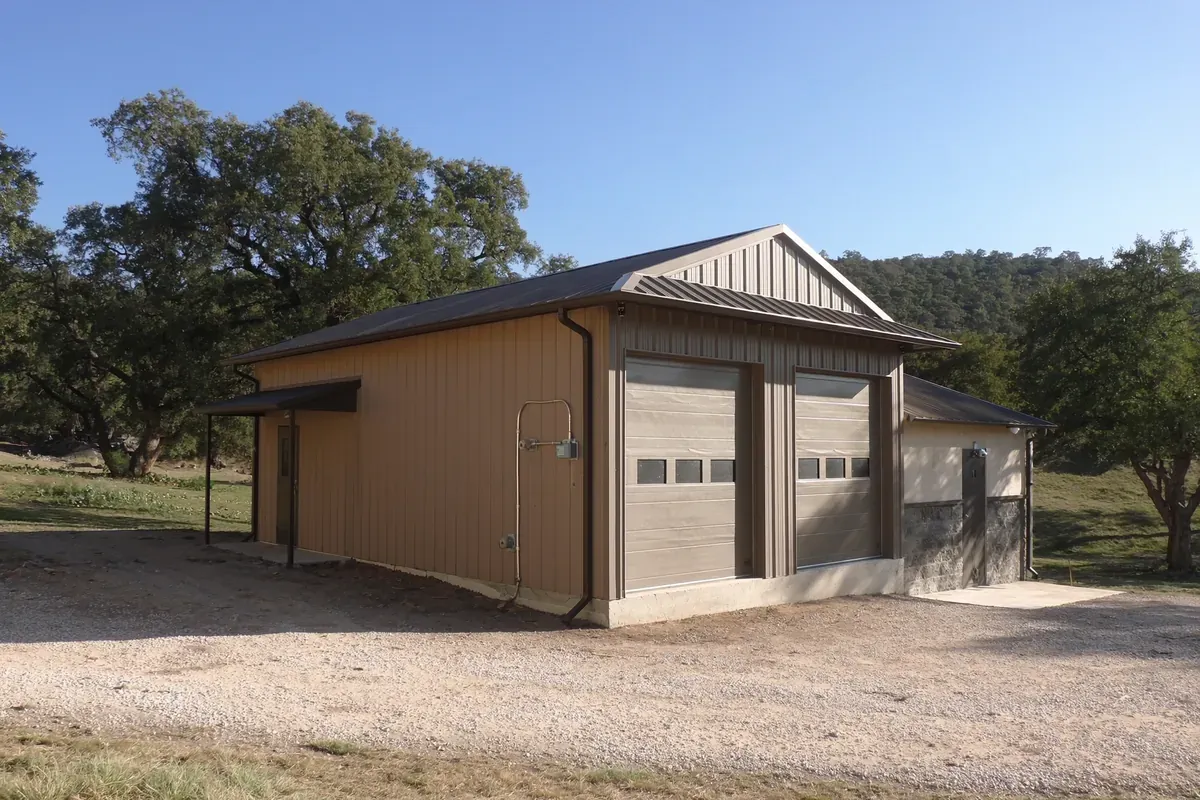 A detached metal workshop and garage with roll-up doors under live oaks in the Texas Hill Country